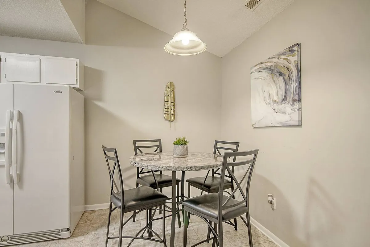 A cozy dining nook with a round dining table set beneath a stylish light fixture, accompanied by artwork and a refrigerator in the background. The arrangement feels intimate and welcoming, ideal for family meals or morning coffee. The proximity to the kitchen reinforces the open-plan layout described in the section.