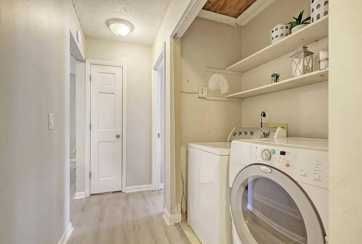 An interior view of a laundry area with a washing machine, open shelves, and doorways leading to other parts of the condo. The space is straightforward and accessible, emphasizing ease of movement.