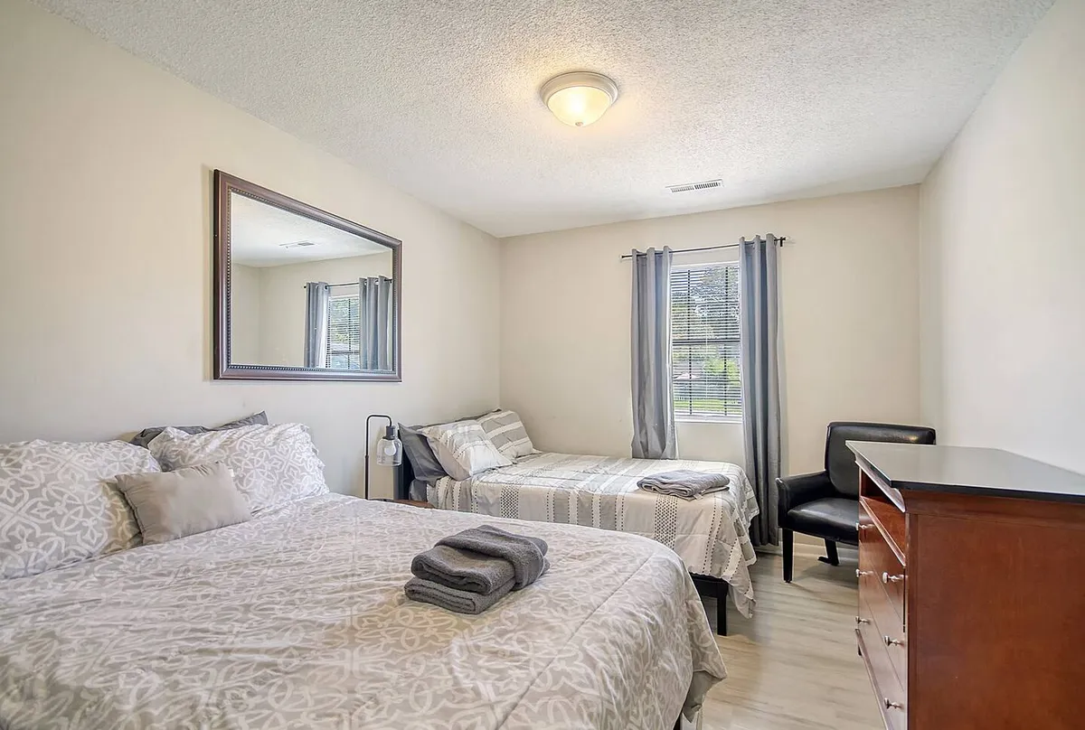 This image presents a bedroom with two beds, a mirror, and a dressing table, set in a comfortable, softly lit environment. The arrangement suggests ample space for relaxing, reading, and enjoying company at the end of the day.