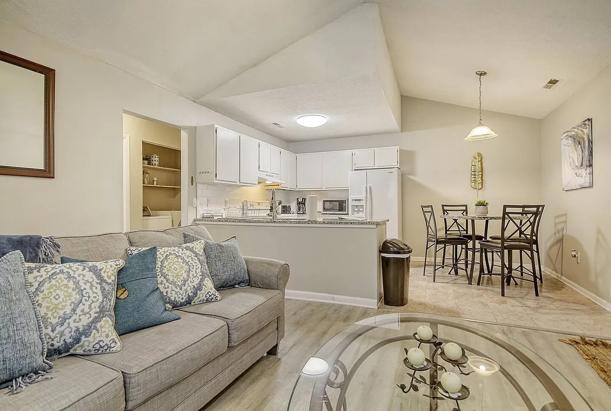 The image showcases a bright, open-plan living area featuring a comfortable sofa with decorative pillows, a stylish dining table set for shared meals, and a modern kitchen fully equipped for cooking. Warm lighting and thoughtfully arranged decor create a cozy, welcoming ambiance.
