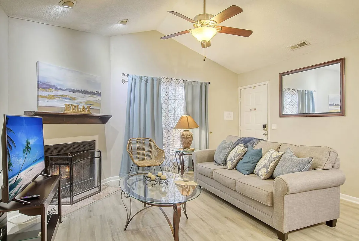 A cozy living room featuring a plush sofa arranged around a sleek coffee table, with a fireplace serving as the focal point. The Smart TV, flanked by elegant curtains, is prominently displayed above the fireplace, inviting guests to relax and enjoy entertainment. Sunlight streams in, enhancing the inviting ambiance and highlighting the open, airy layout.