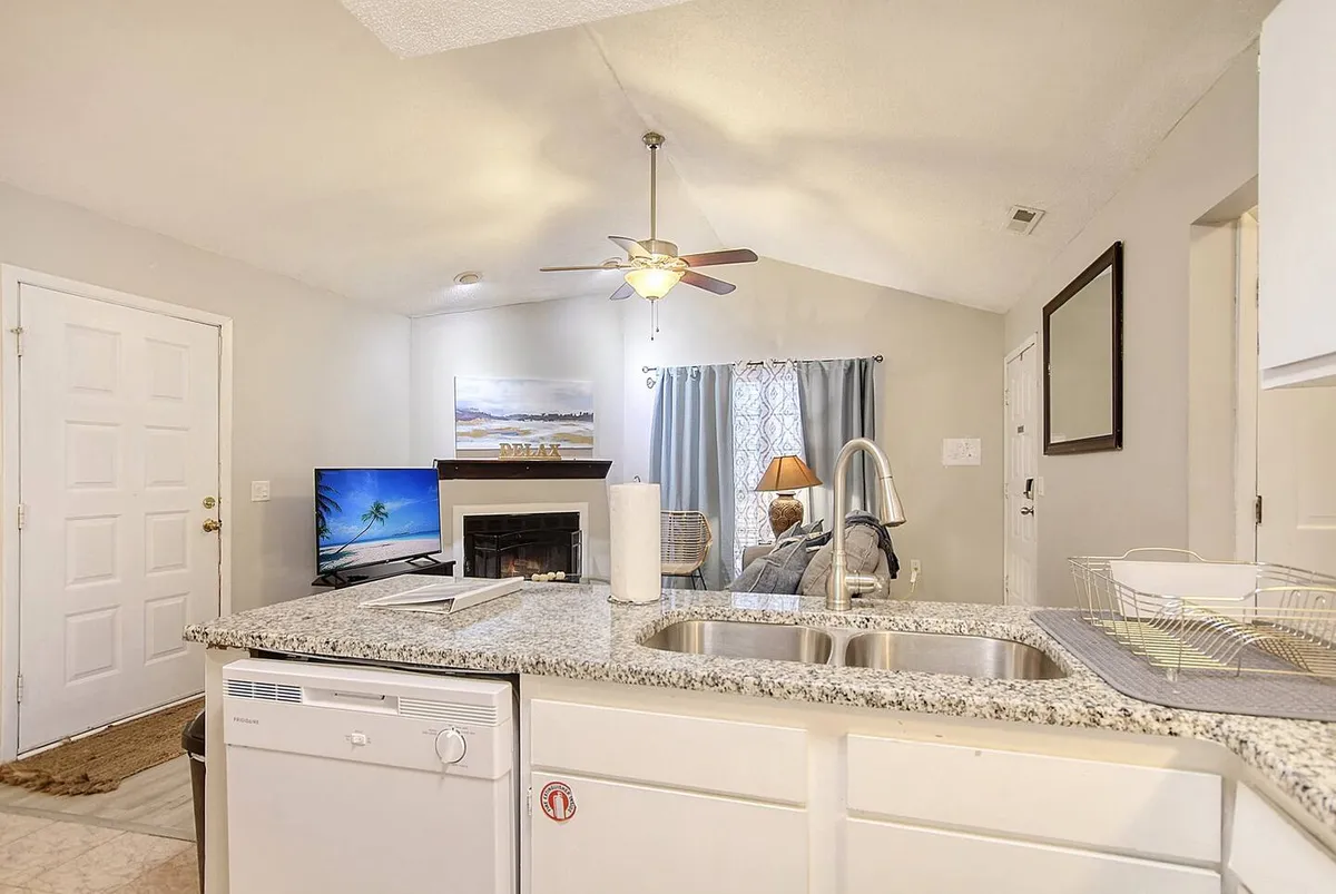A wide shot that captures the open-concept layout, blending the kitchen island with the adjacent living room area. The living space includes a TV and fireplace, flanked by curtains, and is arranged for both relaxation and socializing. The image highlights seamless transitions between spaces, with natural light filling the room and a cozy, inviting mood throughout.